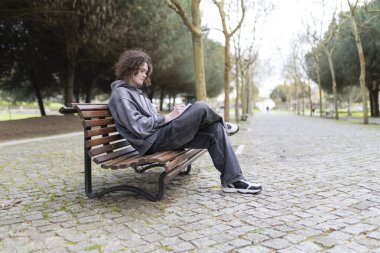 Şehir parkındaki ahşap bankta dinlenirken notlar alan konsantre erkek öğrenci.