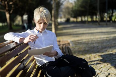İlkokul öğrencisi, bir bankta oturmuş, temiz hava ve güneş ışığının tadını çıkarırken notları inceliyor.