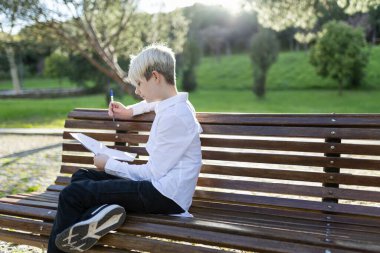 Güneşli bir günde parkta bankta oturan konsantre öğrenci notları yazıyor.