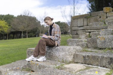 Genç kadın arkeolog araştırmasının notlarını alırken antik harabelerin basamaklarında oturuyor.