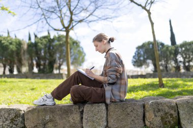 Genç kadın ressam, parkta taş bir çıkıntının üzerinde oturmuş, temiz havanın ve doğal çevrenin tadını çıkarırken bir defterde çizim yapıyor.