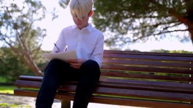Sarışın çocuk bir kağıt parçasına yazı yazıyor. Parktaki bankta oturuyor.
