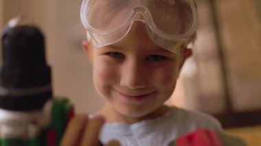 Emniyet gözlüğü takan gururlu genç bir çocuk oyuncak bir elektrikli matkap tutuyor ve gülümsüyor, evde oynuyor.