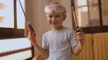 Güvenlik gözlüğü takan gülümseyen çocuk oyuncak testere ve çekiç tutuyor, küçük bir inşaatçı gibi davranıyor.