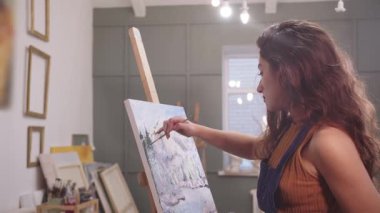 Bir sanat stüdyosunda odaklanmış bir kadın tuvalde fırçayla bir dağ manzarası çiziyor.