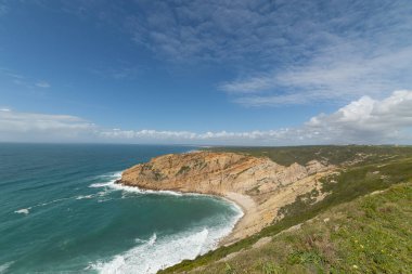 Atlantik okyanusu dalgaları engebeli kayalıklara çarpıyor ve mavi gökyüzünün altında tenha bir kumsal.