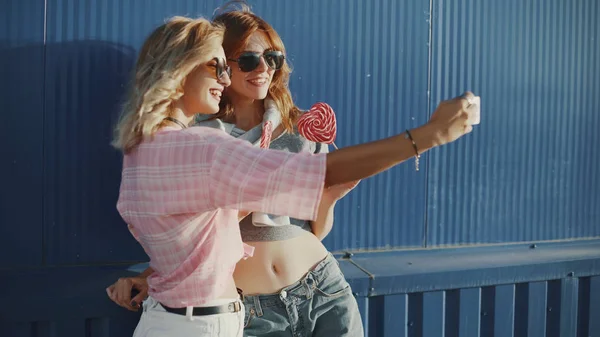 Two young attractive women in sun glasses talking, eat lollipop and take a selfie on street