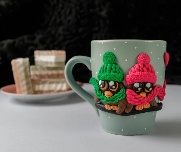 The original mug with decorative figurines made with their own hands. A lovely birthday present. Figures on the cup are fashioned hand made from colored clay. Composition with plate of sweets — Stock Photo, Image