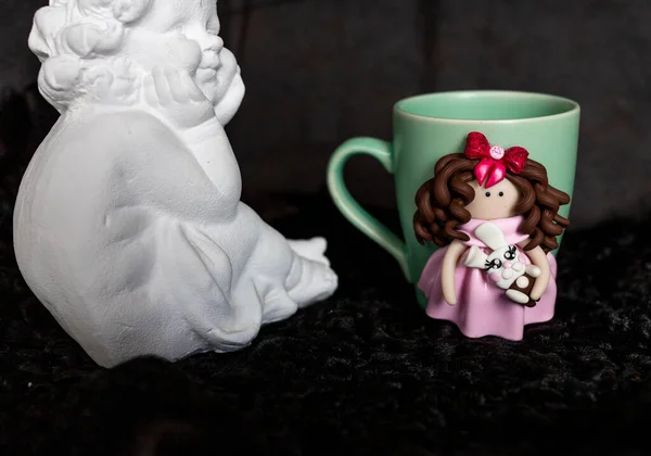 La tasse originale avec des figurines décoratives faites de leurs propres mains. Un joli cadeau d'anniversaire. Les figures sur la tasse sont façonnées à la main à partir d'argile colorée. Composition avec ange en plâtre — Photo