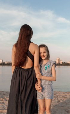 Anne ve kızı gölün kıyısında duruyorlar. Kız kameraya doğru bakıyor ve gülümsüyor ve kadın ters yöne bakıyor. İkisinin de uzun saçları var..