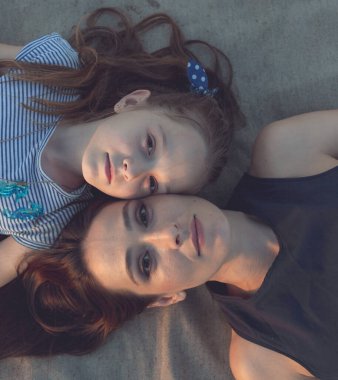 Anne ve kızı kum renkli bir paspasın üzerinde dinleniyorlar. Kamera onlara yukarıdan, omuzlarından bakıyor. Kadın ve kız çok güzel, Avrupa görünüşlü.