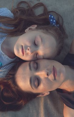 Anne ve kızı kum renkli bir paspasın üzerinde dinleniyorlar. Kamera onlara yukarıdan, omuzlarından bakıyor. Kadın ve kız çok güzel, Avrupa görünüşlü.