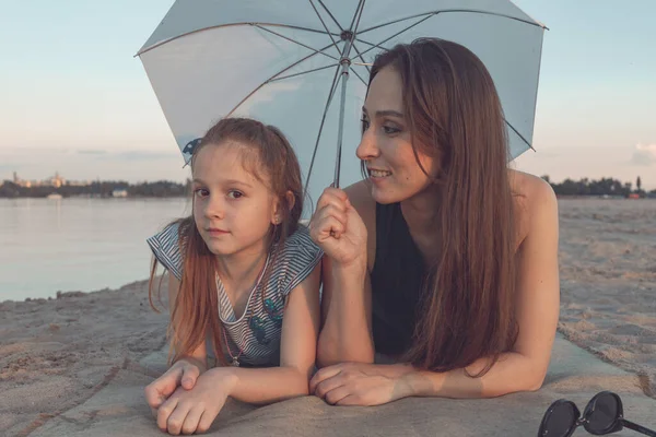 Kız ve annesi kumsalda bir Mat 'ta beyaz bir şemsiyenin altında uzanıyorlar ve mutlu bir şekilde gülümsüyorlar. Suyu onlardan uzakta görebilirsin..