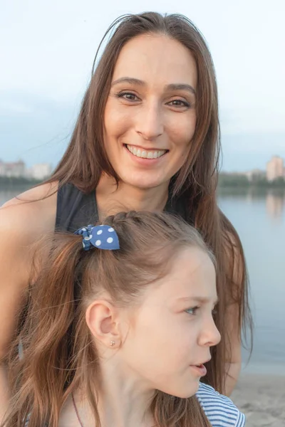Mom and daughter in front of the camera. Woman smiles beautifully and looks at us. Girl is very emotional. Real people. Background - lake, sky and forest
