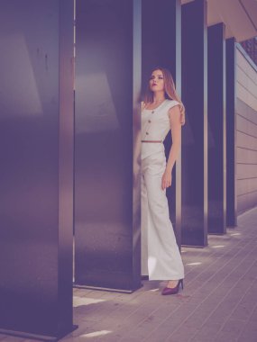 Şehir binasının arka planında poz veren güzel beyaz bir kız. Kız beyaz bir takım elbise giyiyor. Çok güzel saçları ve makyajı var.