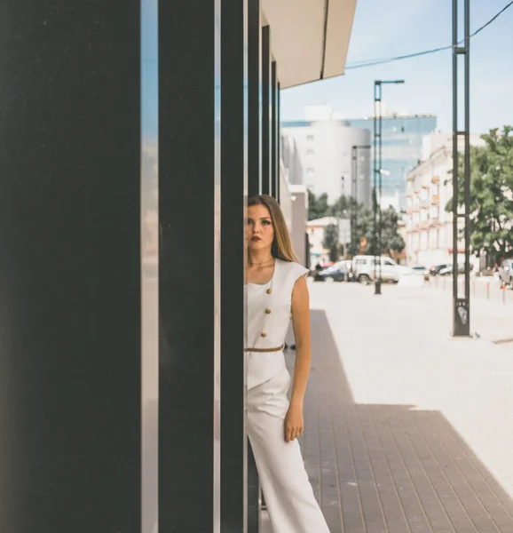 Uma linda menina branca posando no fundo de um prédio da cidade. A menina está vestida com um terno branco. Ela tem cabelo lindo e maquiagem — Fotografia de Stock