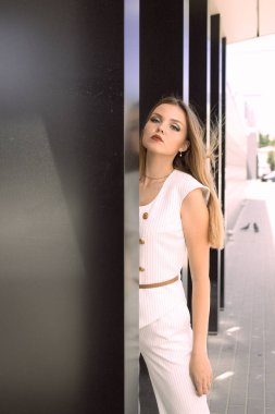 Şehir binasının arka planında poz veren güzel beyaz bir kız. Kız beyaz bir takım elbise giyiyor. Çok güzel saçları ve makyajı var.