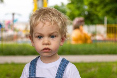 Mavi tulumlu ve beyaz tişörtlü iki yıllık küçük bir çocuk parkta yürüyor ve etrafında olup biten her şeye şaşırıyor.