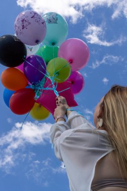 Beyaz, bol elbiseli, sarı saçlı bir kadın bir sürü balon tutuyor. Sonsuz mavi gökyüzü ve bulutlar.