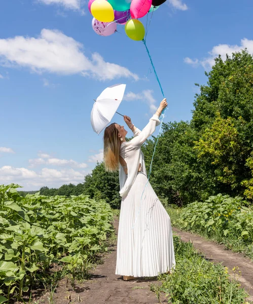 Eine Frau im weißen, lockeren Kleid und mit blonden Haaren hält einen ganzen Strauß Luftballons in der Hand. Der grenzenlose blaue Himmel und Wolken. Stockfoto