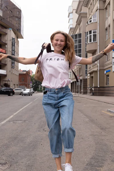 Uma linda menina sorridente em uma camiseta branca e jeans posa nas ruas da cidade. Ela tem cabelo comprido e não tem maquilhagem. a arquitetura da cidade velha é um pano de fundo em torno dele — Fotografia de Stock