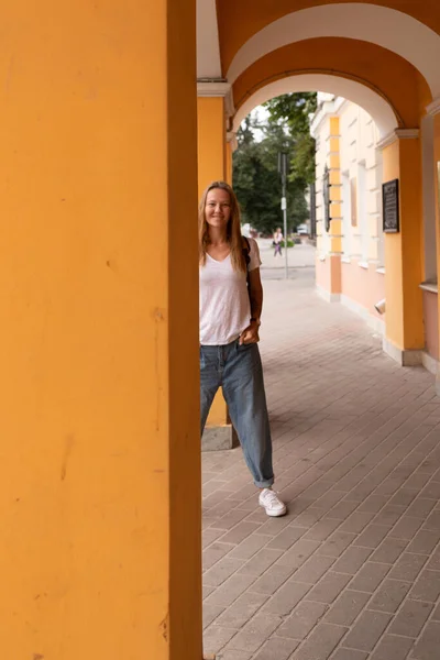 En vacker leende flicka i vit t-shirt och jeans poserar på gatorna i staden. Hon har långt hår och inget smink. arkitekturen i den gamla staden är en bakgrund runt den — Stockfoto