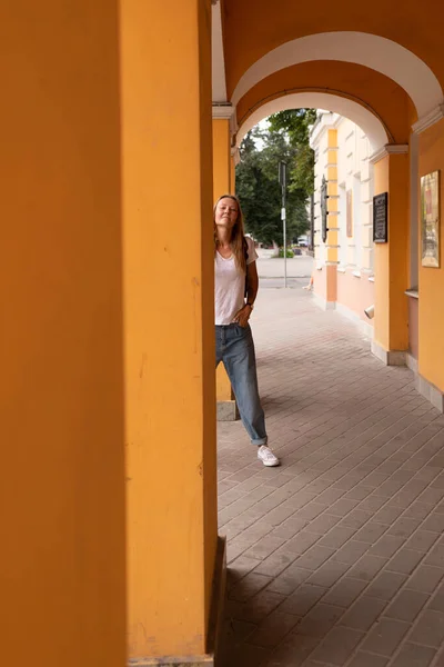 Krásná usměvavá dívka v bílém tričku a džínách pózuje na ulicích města. má dlouhé vlasy a žádný make-up. architektura starého města je kulisou kolem něj — Stock fotografie