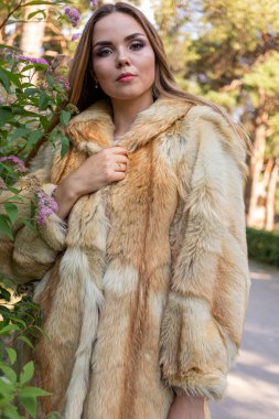 Uzun saçlı ve anlamlı gözlü güzel bir kız parkta kürk giymiş poz veriyor.