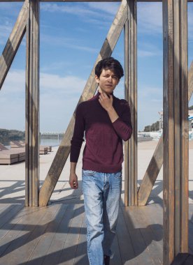 Doğu görünümlü, siyah saçlı, yakışıklı ve etkileyici yüzlü genç bir adam ağaçların ve mimarinin arka planına karşı parkta poz veriyor.