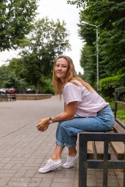 Jovem bela mulher elegante sentada na rua em um dia ensolarado de verão. Uma menina da moda usa uma tshirt branca e jeans azuis. Uma mulher com cabelos longos e lisos e tatuagem — Fotografia de Stock