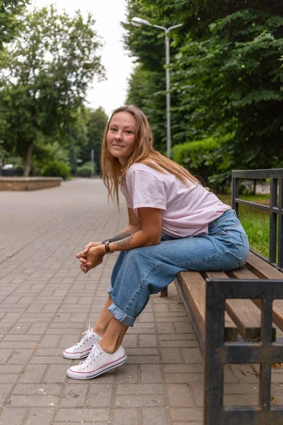 Jovem bela mulher elegante sentada na rua em um dia ensolarado de verão. Uma menina da moda usa uma tshirt branca e jeans azuis. Uma mulher com cabelos longos e lisos e tatuagem — Fotografia de Stock