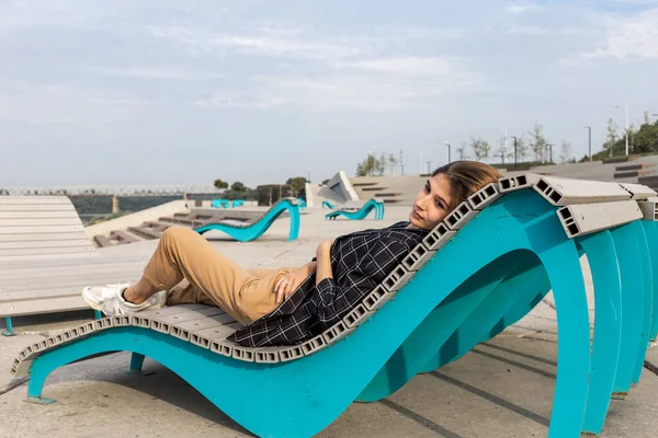 Genç ve güzel bir kadın güneşli bir yaz gününde bankta oturuyor ve kameraya bakıyor. Uzun düz saçlı bir kadın.