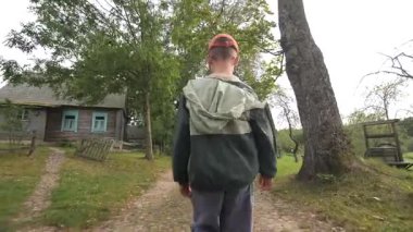 Büyüleyici ahşap evler ve yemyeşil ağaçlarla çevrili, dolambaçlı bir köyde dolambaçlı bir yolda yürüyen genç bir çocuk, huzurlu bir kırsal bölgede huzur içinde.