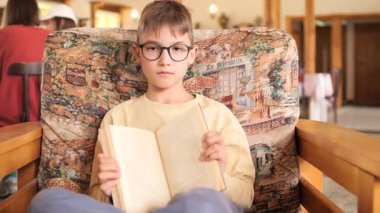 Gözlüklü, koltukta oturmuş kitap okuyan, rahat bir ortamda edebiyat yapan, muhtemelen kütüphane ya da oturma odası olan zeki bir çocuk.