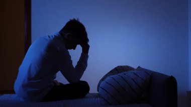 Elleri havada yatakta oturan bir adam, zihinsel sağlık sorunları yüzünden stres ve endişe yaşıyor ve odasında karanlık bir gecede uykusuzluk çekiyor.