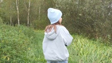 Açık mavi bir şapka ve ceket giyen genç bir kız güneşli bir ormanda çimenli bir yolda yürüyor, doğanın huzurunun tadını çıkarıyor ve ormanın güzelliğini keşfediyor.
