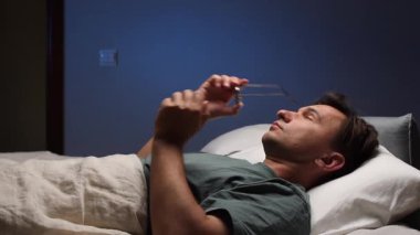 Bir adam yatağa gitmeden önce gözlüğünü çıkarır ve yorgun gözlerini ovuşturur. Görme duyusunda bozulma. Gün boyunca stres ve yorgunluk birikmiş. Uykusuz bir adam gece yatağında uzanıyor, karanlık bir yatak odasında yorgun gözlerini ovuyor, uykusuzlukla boğuşuyor.. 