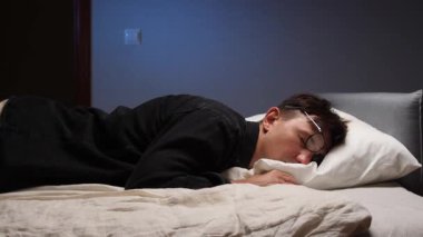 Yorgun bir adam gözlükleriyle bir yatakta uyuyor, muhtemelen uzun bir çalışma ve çalışma gününden sonra, yorgunluk, dinlenme ve modern hayatın talepleri üzerine,