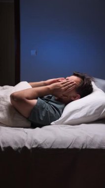 vertical video Sleepless man lying in bed at night covering his ears with his hands, trying to fall asleep in his bedroom, suffering from insomnia and sleep disorder
