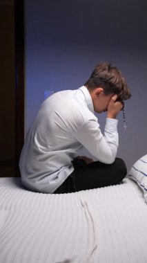 vertical video christian man sitting on a bed in a bedroom, holding a rosary and covering his face with his hand, reflecting emotions of sadness, loneliness, and repentance during a moment of private prayer