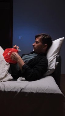 vertical video Sick man is lying in bed at night, covered with a blanket, and opening a first aid kit with a worried expression, searching for medicine or treatment