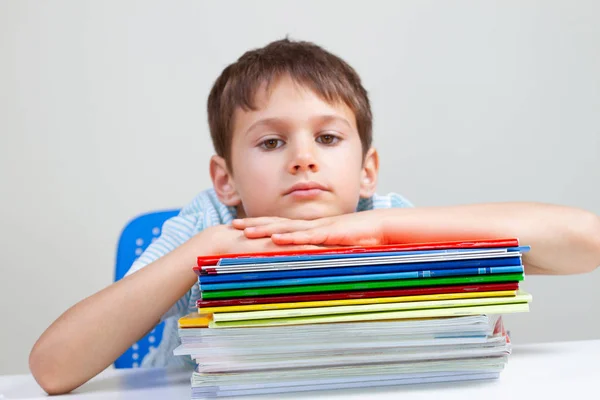 Scolaretto sconvolto seduto alla scrivania con mucchio di libri scolastici e quaderni — Foto Stock