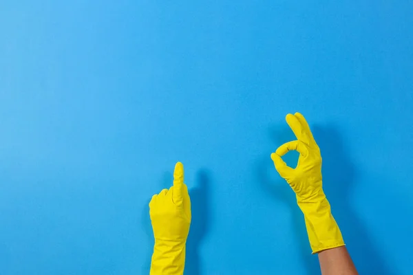 Mãos de mulher com luvas de borracha amarela fazendo um gesto que significa ok e aponta para cima com o dedo, fundo azul — Fotografia de Stock