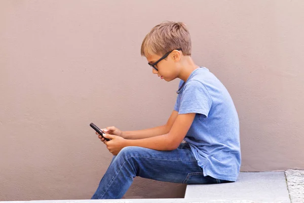 Menino com smartphone sentado nas escadas ao ar livre. Kid wathing to screen, ler, digitar, jogar jogos — Fotografia de Stock