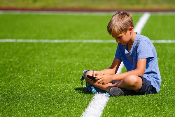 Boy sitting with cell phone sitting in empty football stadium field and looking video, typing, playing