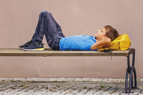 Niño con mochila bajo la cabeza acostado en el banco al aire libre — Foto de Stock