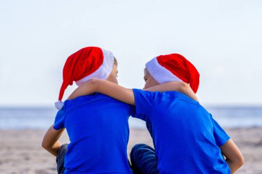 Noel tatili. Çocuklar güneşli kumsalda sarılma kırmızı Santa şapkalar Sit