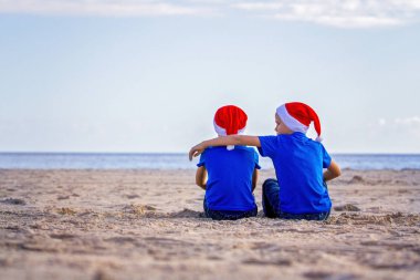 Noel tatili. Çocuklar güneşli kumsalda sarılma kırmızı Santa şapkalar Sit