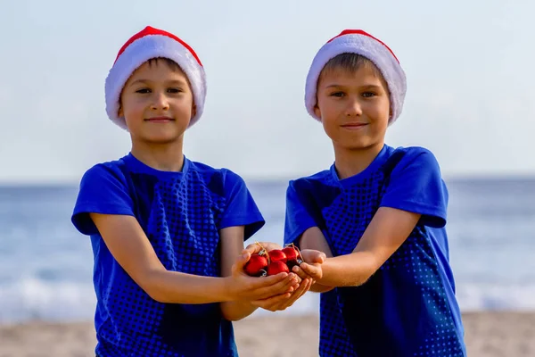 Vacaciones de Navidad. Niños en sombreros rojos de Santa celebración de la decoración de Navidad en la playa soleada —  Fotos de Stock