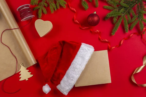 Készül a karácsony: csomagolópapír, dekorációk és jelen díszdobozban, Mikulás kalap, piros háttér — Stock Fotó